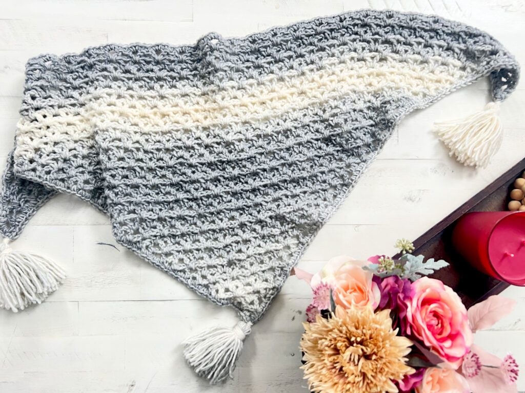 A crochet triangular shawl with a gradient of grey to white tones, featuring a textured pattern and tassels on each corner, is laid out on a light wooden surface. To the right, there is a tray with a red candle and a bouquet of pink and cream flowers. -Marly Bird