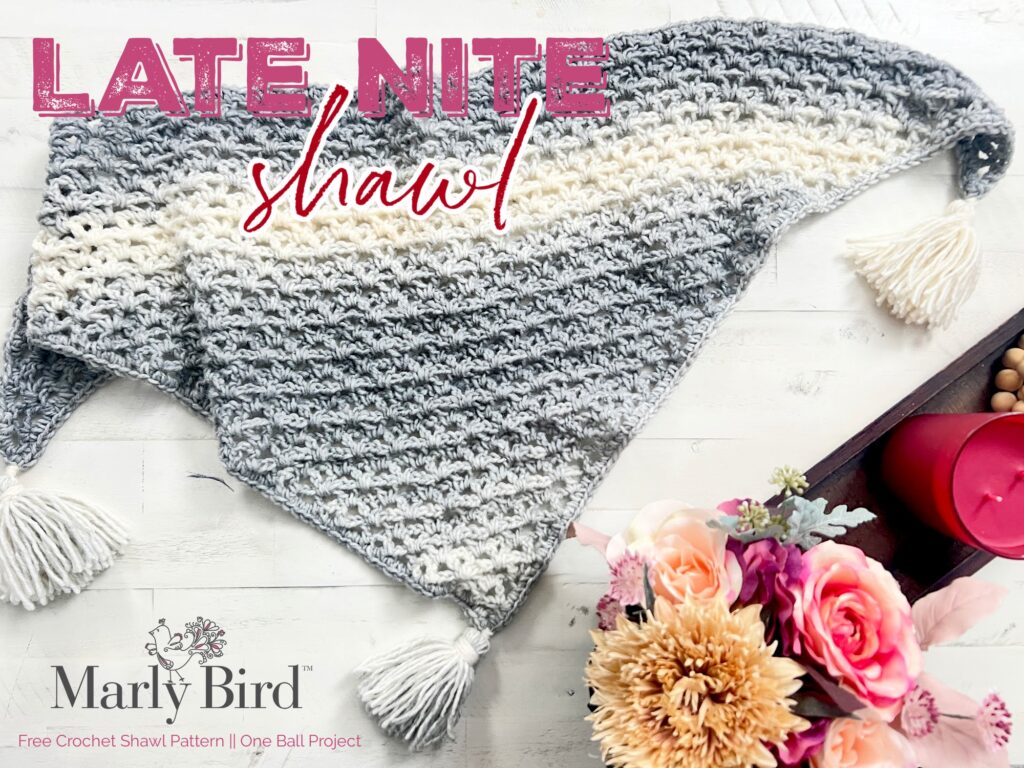 A crochet shawl with a gradient pattern ranging from off-white to various shades of gray is laid flat on a white surface. It has tassels on three corners. Nearby are a pink candle, pink flowers, and a "Marly Bird" logo with "Late Nite Shawl" text above. -Marly Bird