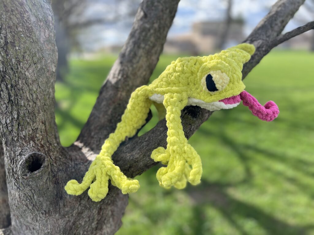 A bright green, crocheted frog with long limbs and big eyes hangs on a tree branch. This leggy frog, created from a delightful crochet pattern, has a pink protruding tongue and is set against a backdrop of a green grassy area with another tree, under a partly cloudy sky. -Marly Bird