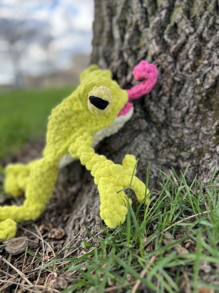 A Leggy Frog Crochet Pattern comes to life with a yellow frog featuring white eyes and a pink tongue, positioned as if it's climbing up a tree trunk. The charming scene is set on a grassy patch with visible tree roots, complemented by a blurred background. -Marly Bird