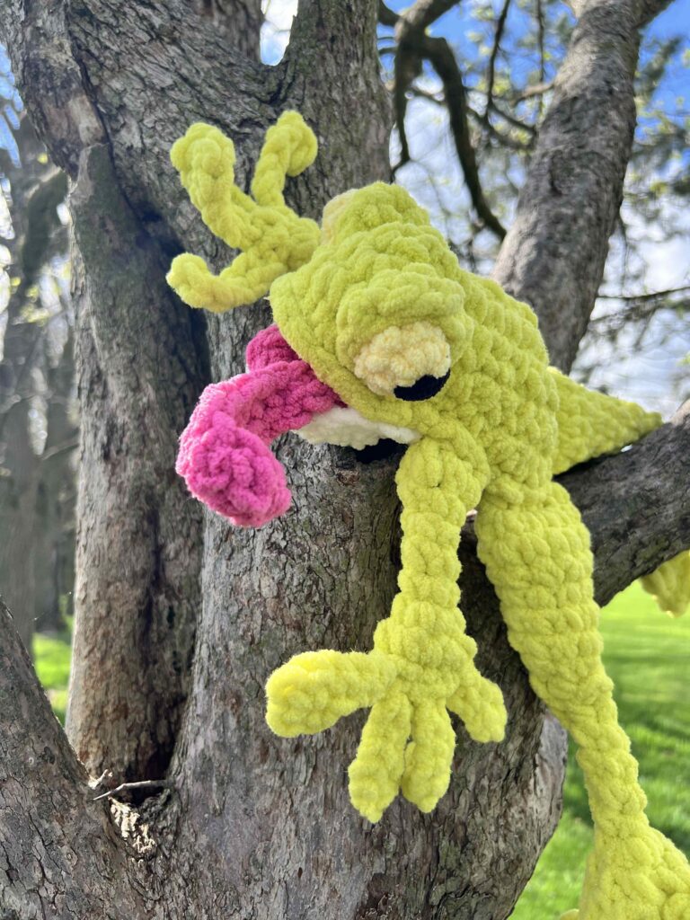 A leggy plush yellow frog with a pink tongue and bulging eyes is draped over a tree branch in a sunlit outdoor setting, with grass and other trees visible in the background. The crochet pattern gives the handmade toy its textured fabric, enhancing its playful appearance. -Marly Bird