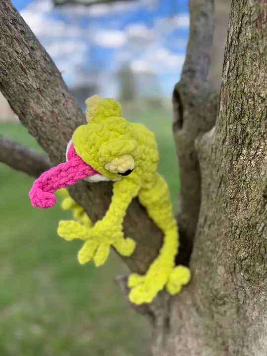 A crocheted green and yellow chameleon with a long pink tongue is draped over a tree branch outdoors, inspired by free knitting and crochet patterns, with a blurred background of grass and blue sky. -Marly Bird