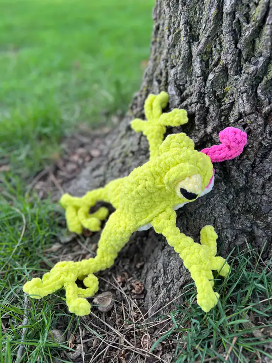 Amigurumi frog plush in bright yellow yarn with a pink tongue, shown climbing a tree base on grass; visible crochet texture.