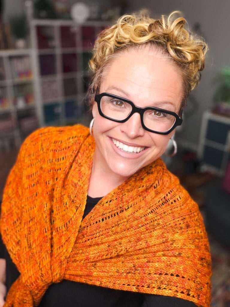 A person models a vibrant, textured Fire Sprite Shawl; knit stitches and bright colorwork are visible against bookshelves.