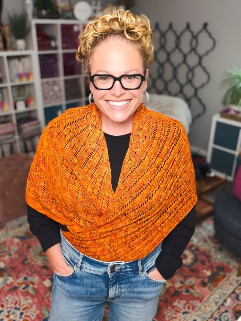 A Lehabah Fire Sprite Shawl drapes over a black top, showing intricate knit stitches and vibrant colors in a cozy craft room setting.