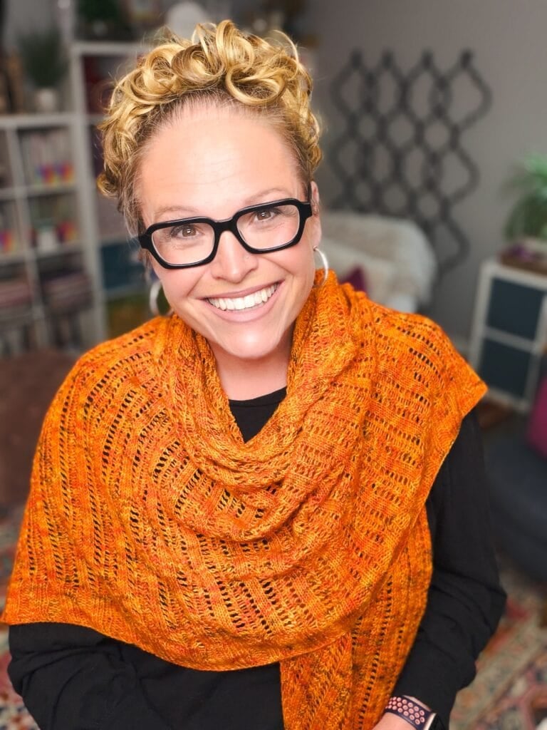 Person wearing a bright orange knitted Lehabah Fire Sprite Shawl with eyelet stitch detail over a black top in a colorful room.