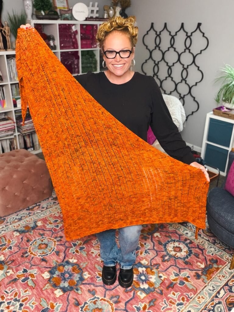 An orange Lehabah Fire Sprite Shawl with textured stitches is displayed by a person in a colorful, book-filled room.