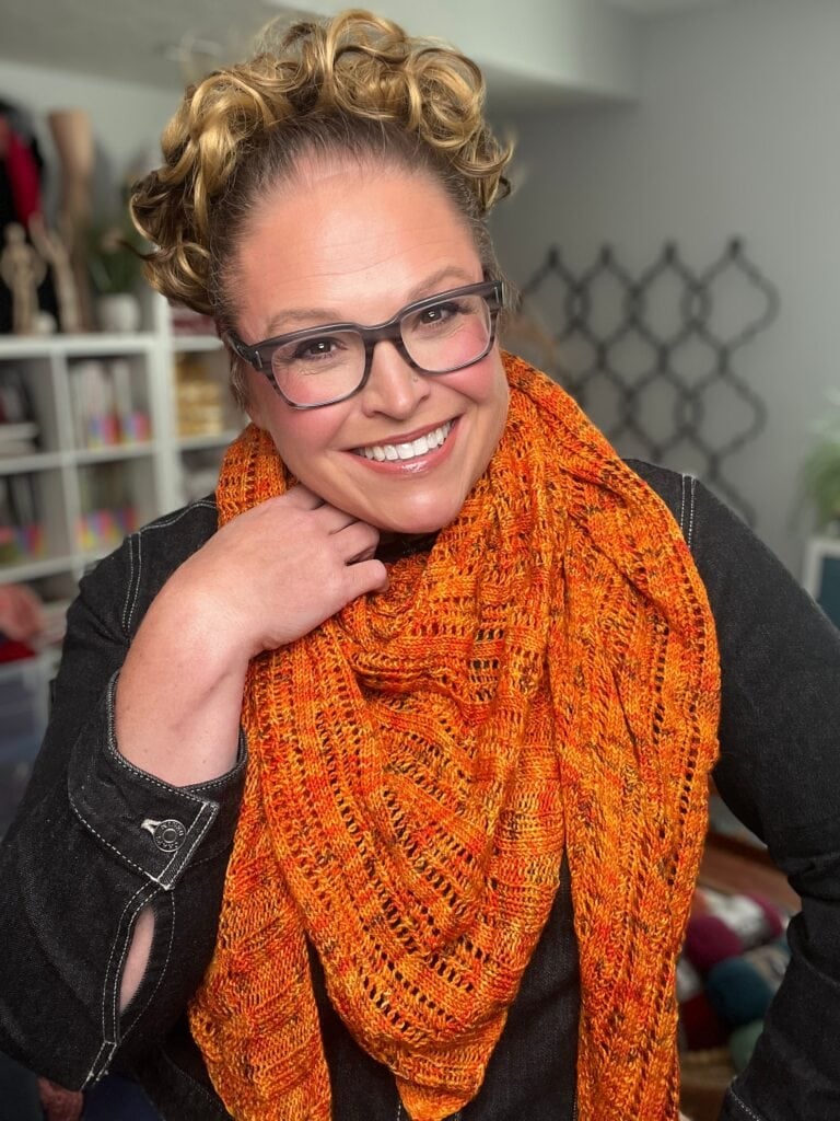 A person wears a textured, bright orange knitted scarf styled like the Lehabah Fire Sprite Shawl, with yarn shelves in the background.