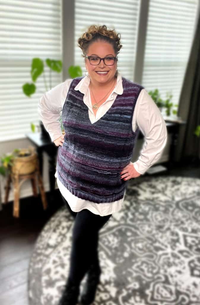 A person with curly hair and glasses is smiling, standing on a patterned rug in a room with plants. They are wearing a white shirt under a knit vest in purple and gray, which perfectly shapes the shoulders, paired with black pants. -Marly Bird