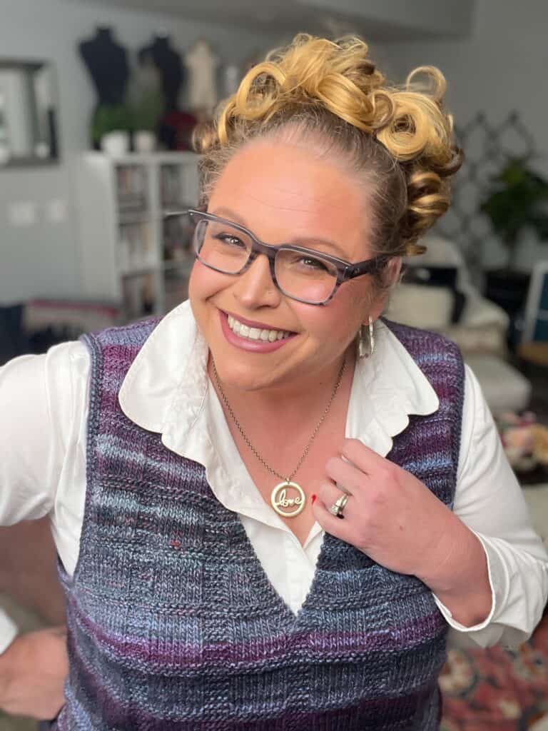 A smiling person with curly hair, wearing glasses and a cozy knit vest over a white shirt, poses indoors. They proudly display a necklace with a "love" pendant. The background features bookshelves lined with plants, adding warmth to the scene. -Marly Bird