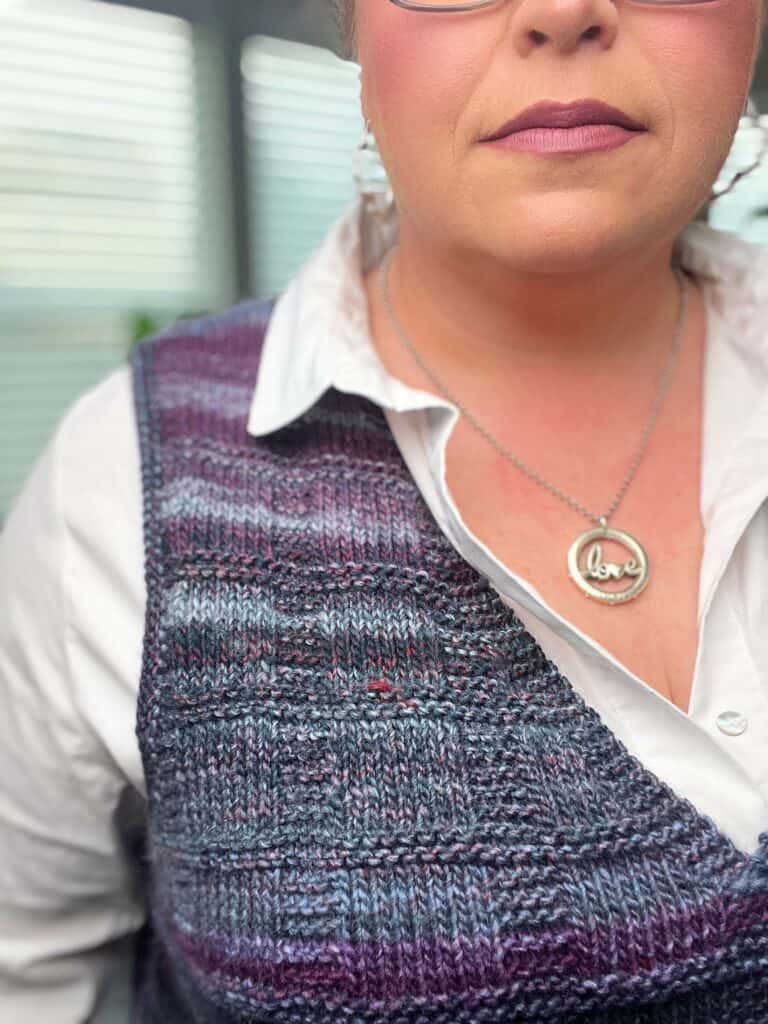 A person wearing a purple and gray knit vest with stylish armholes over a white shirt, accessorized with hoop earrings and a necklace with a round pendant inscribed with "love." The background is softly blurred. -Marly Bird