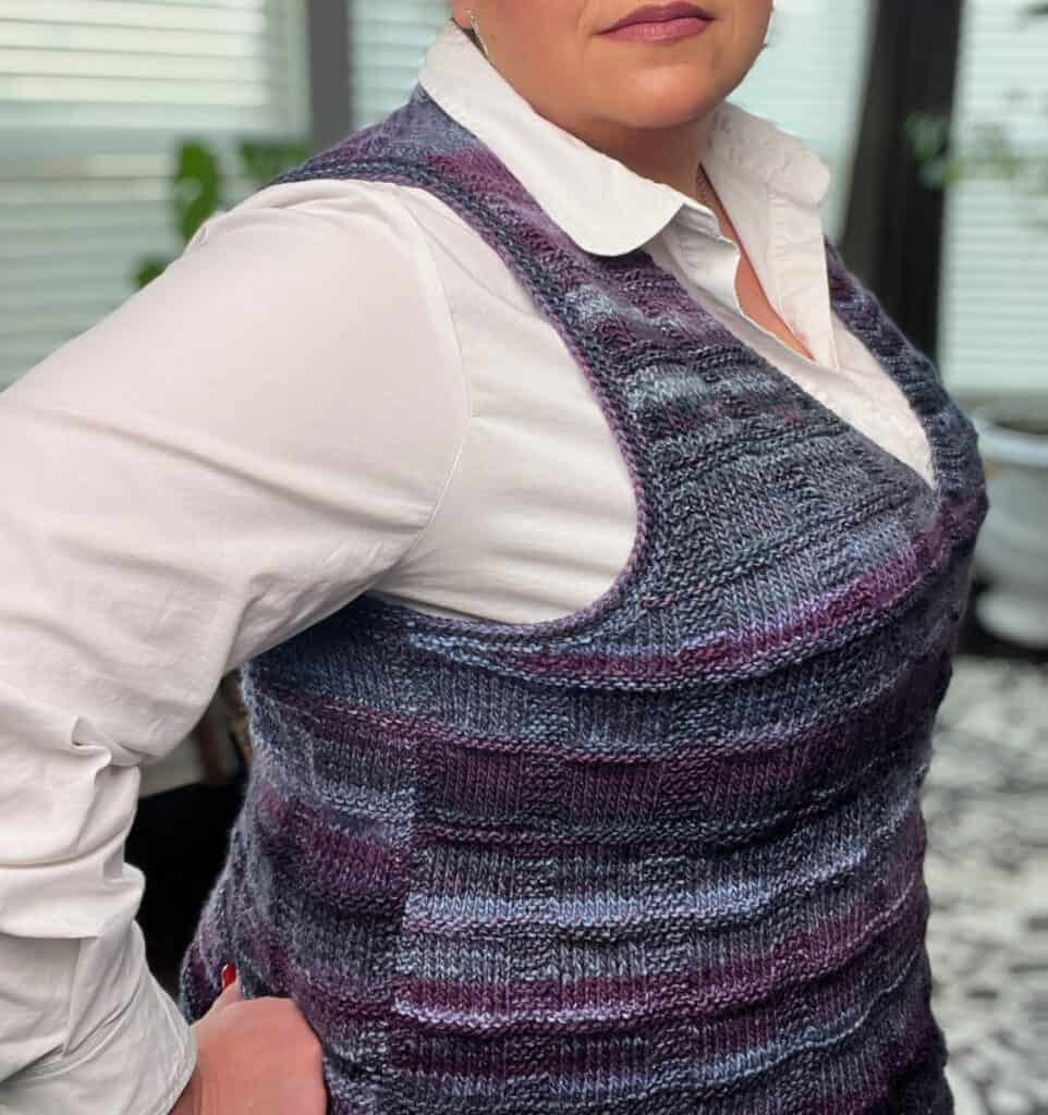 A person wearing a purple and blue knit vest with stylish pockets over a white button-up shirt stands confidently with hand on hip. They're indoors, framed by blurred windows and a plant in the background. -Marly Bird