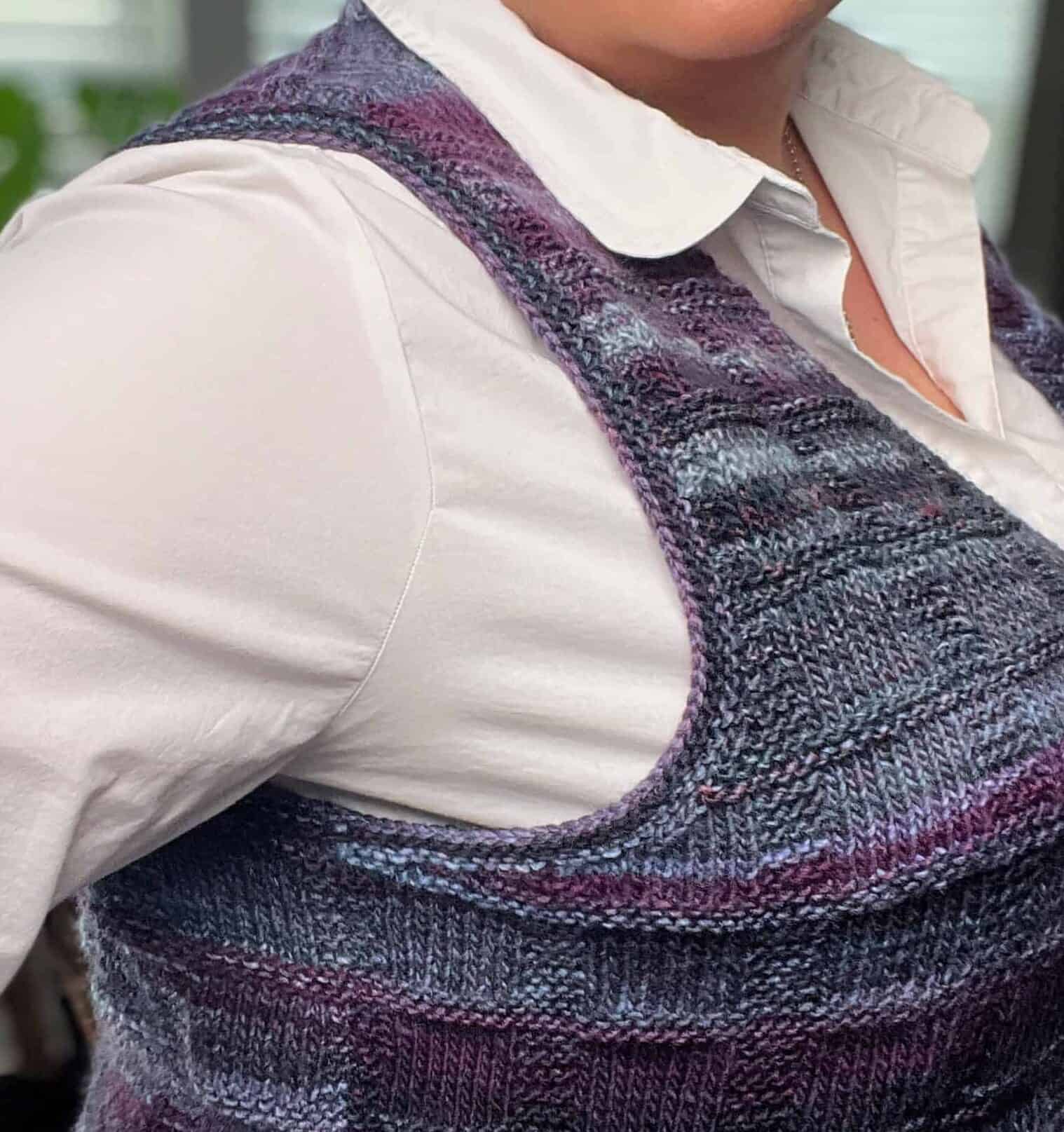 A person wearing a purple and blue knit vest with stylish pockets over a white button-up shirt stands confidently with hand on hip. They're indoors, framed by blurred windows and a plant in the background. -Marly Bird