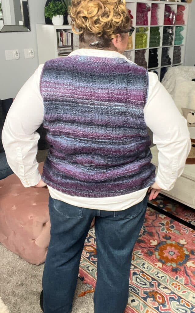 A person with curly hair stands with their back to the camera, showcasing the knit vest's intricate neckline. They're wearing a white shirt beneath, and shelves of colorful yarn enhance the cozy atmosphere. -Marly Bird