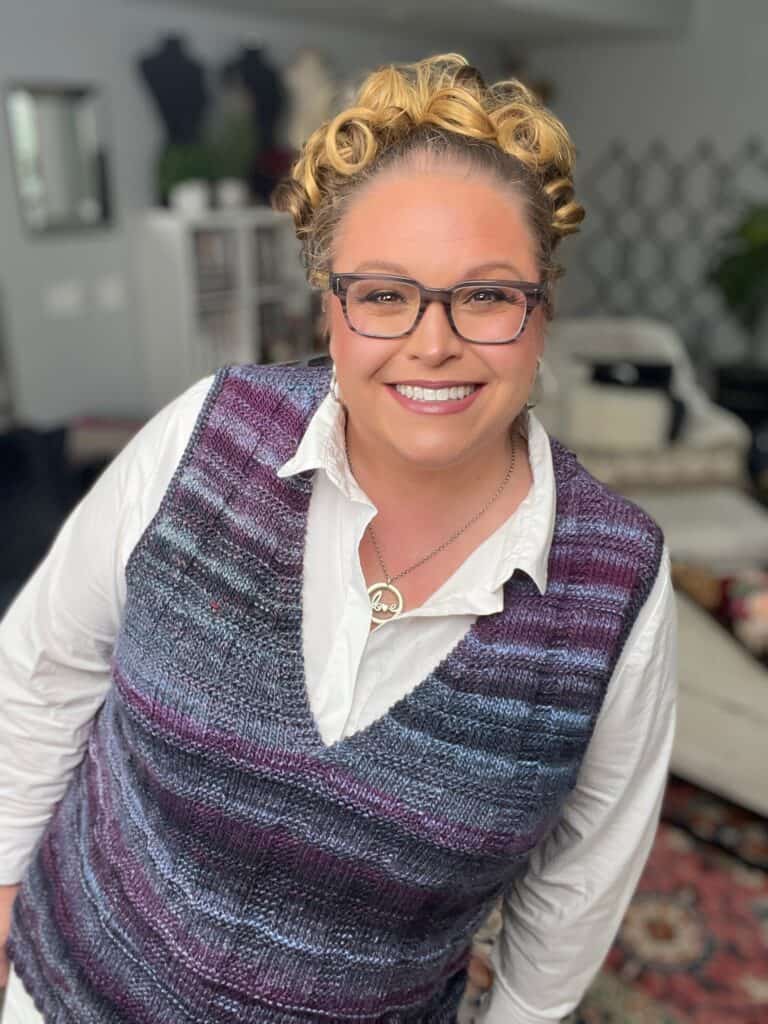 A smiling person in glasses and a white shirt showcases a multicolored knit vest in purple and blue, featuring intricate neckline details. They have curly hair and stand indoors with blurred shelves and a rug behind them. -Marly Bird