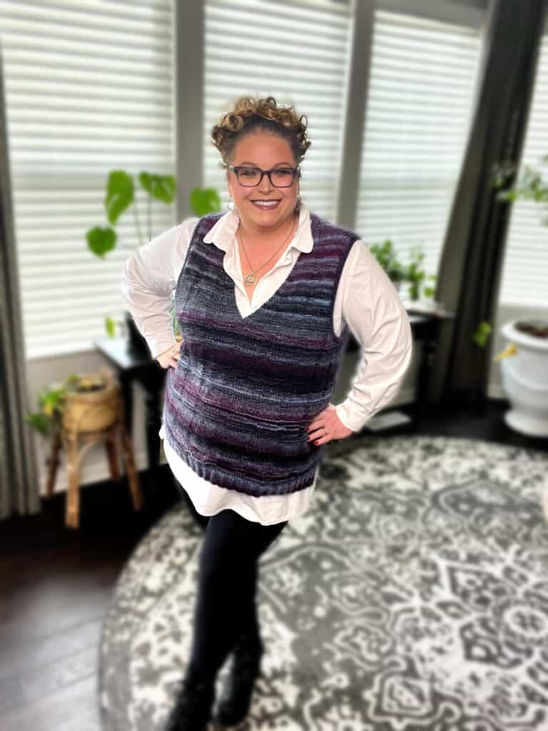 A person smiling indoors with hands on hips, dressed in glasses and a purple striped knit vest over a white shirt. The room boasts a patterned rug, large windows with blinds, and several green plants. -Marly Bird