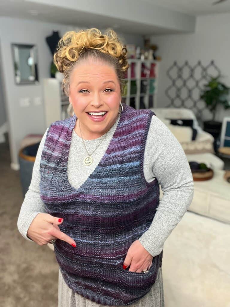 A person with curly hair smiles brightly while pointing at the stylish pocket of their dark striped knit vest. They are wearing a light gray long-sleeve shirt underneath. The background features a cozy living room with a couch and decor. -Marly Bird