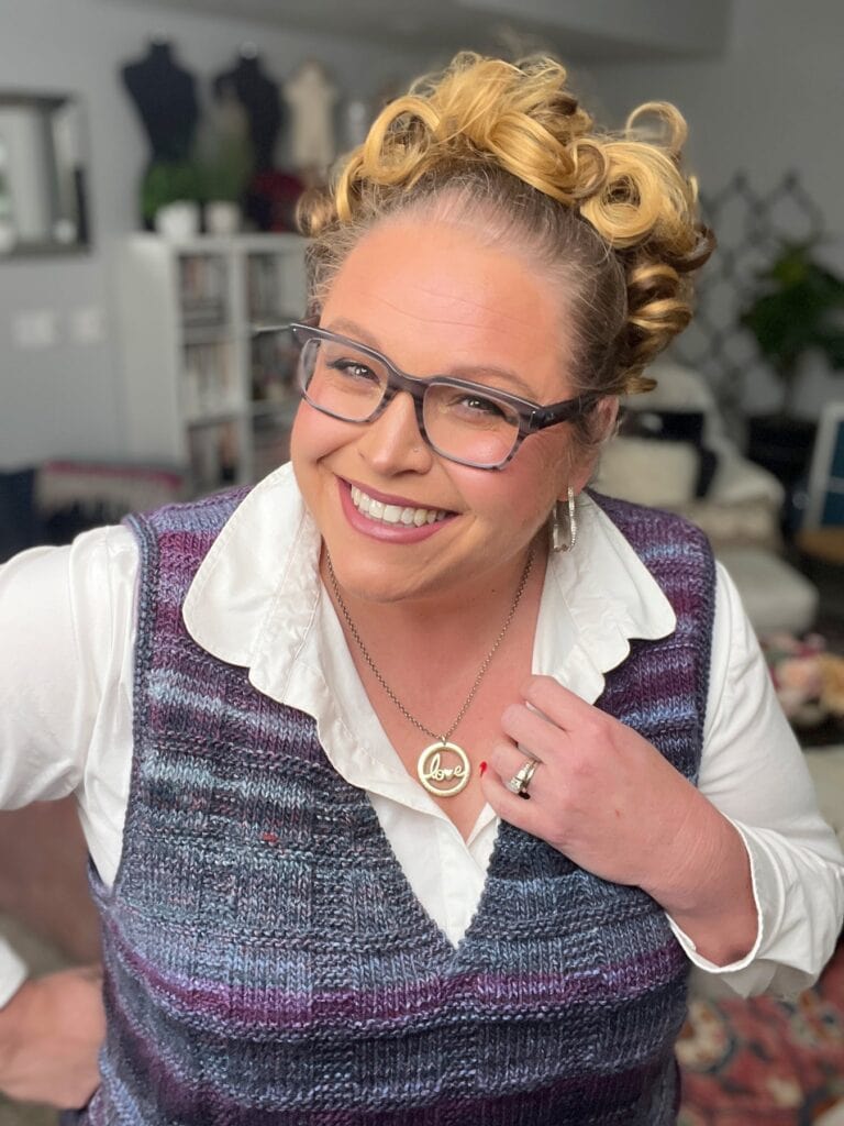 A smiling person with curly blonde hair, glasses, and a purple plaid vest over a white shirt stands indoors. Surrounded by bookshelves and home décor, they showcase their love necklace and ring—perhaps searching for free knitting and crochet patterns. -Marly Bird