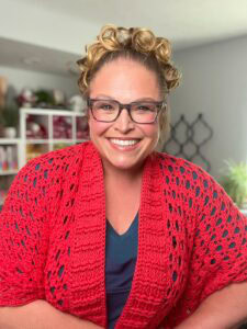 A woman models a bright red, crocheted shawl with visible stitch detail; bookshelves and plants in the cozy background.