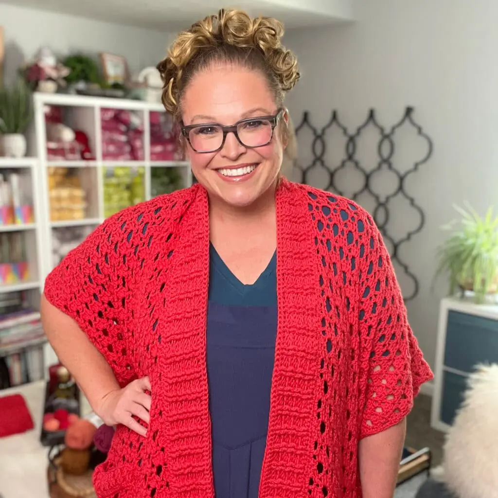 Woman models a chunky, hand-crocheted red cardigan with textured stitches; yarn and plants on shelves in cozy studio background.