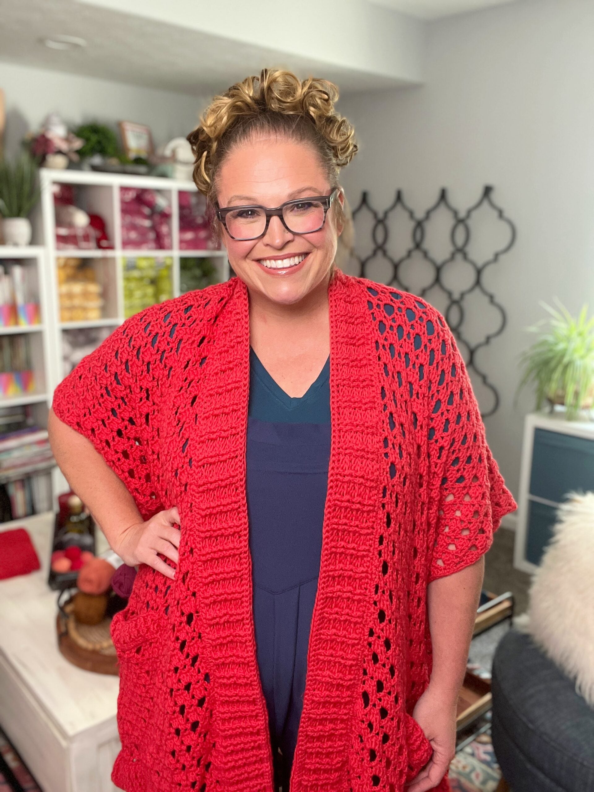 A woman models a red Tunisian crochet shawl, showing textured stitches; bookshelves and plants are visible behind her.