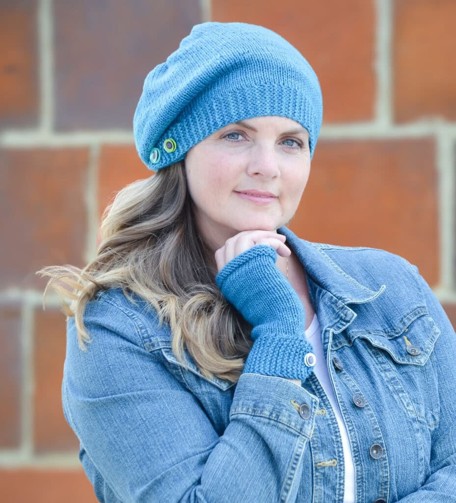 Blue knit hat and fingerless gloves set, featuring textured stitches, modeled with a denim jacket against a brick wall.