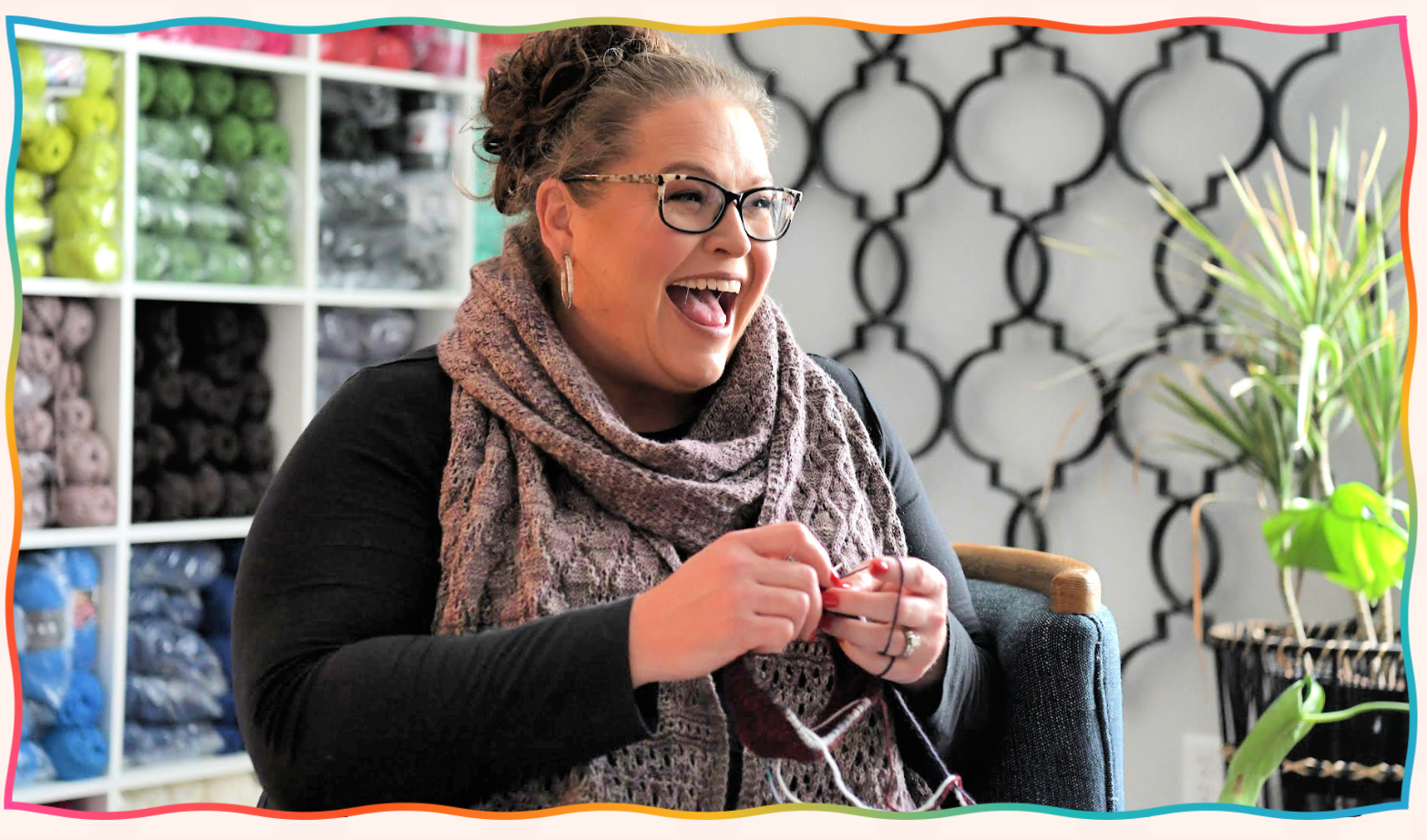 A person with glasses is sitting and knitting, smiling beneath their cozy scarf. In a room adorned with yarn shelves and a plant, they channel their inner Marly Bird. The decorative wall with circular patterns adds an artistic touch to this creative haven. -Marly Bird