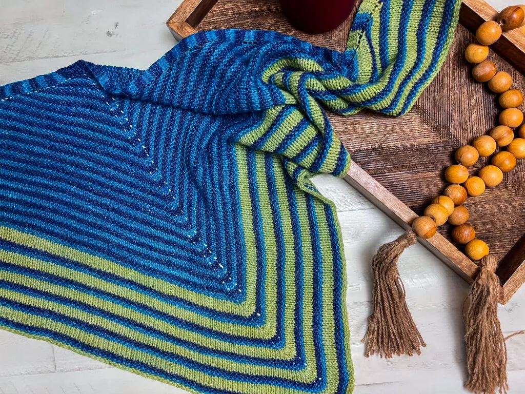 An Easy Knit Triangle Shawl with blue and green stripes is draped on a wooden tray beside a string of wooden beads with tassels. The tray and shawl are displayed on a light-colored surface. -Marly Bird