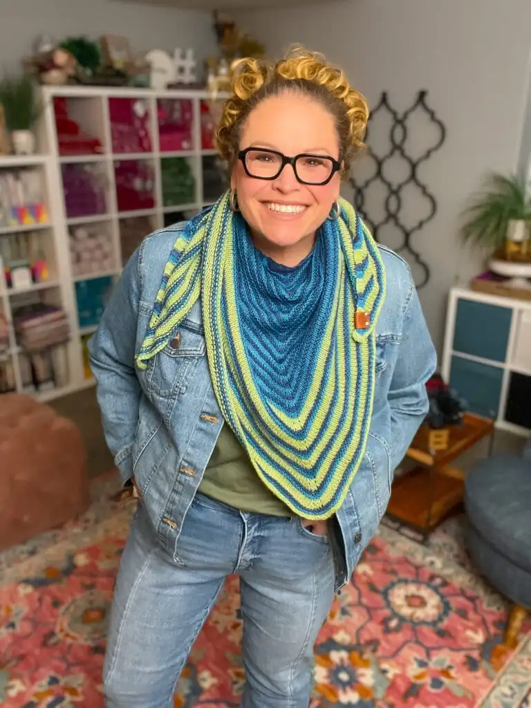 A smiling person with curly blonde hair and glasses wears an Easy Knit Triangle Shawl in blue and green stripes over a denim jacket and jeans, standing in a cozy, colorful room with bookshelves and a patterned rug. -Marly Bird