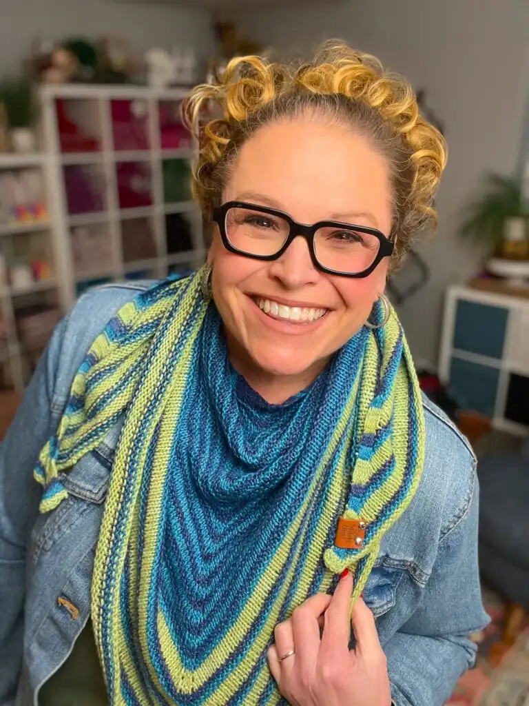 A smiling person with curly blonde hair and black glasses wears an Easy Knit Triangle Shawl in blue and yellow stripes over a denim jacket, standing indoors with shelves and plants in the background. -Marly Bird