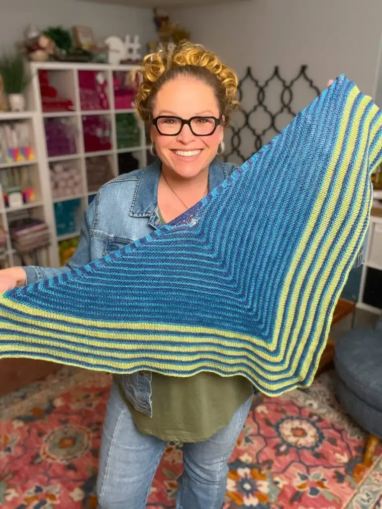 A smiling person with glasses and curly hair holds up an Easy Knit Triangle Shawl in blue, green, and yellow stripes, standing in a cozy, colorful room with shelves of yarn in the background. -Marly Bird