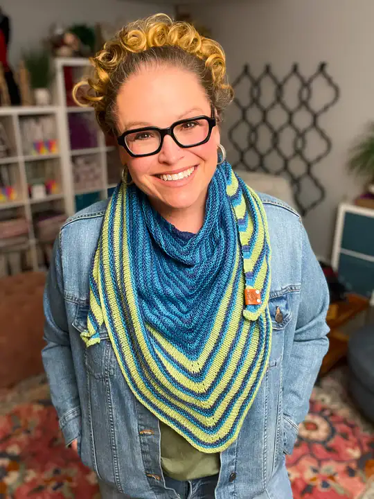 A smiling woman with curly hair and glasses wears a blue and green striped knit shawl made from free knitting and crochet patterns, standing in a cozy, colorful room with shelves and patterned decor in the background. -Marly Bird