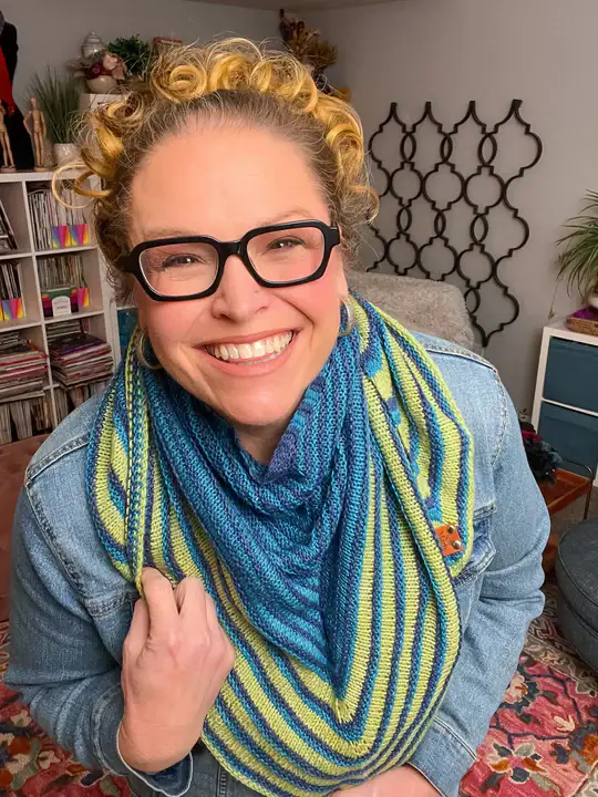 A smiling person with curly blond hair, black glasses, and a denim jacket wears a blue and green striped knitted scarf made from free knitting and crochet patterns. They stand indoors in a cozy, colorful room with plants and decorative items in the background. -Marly Bird