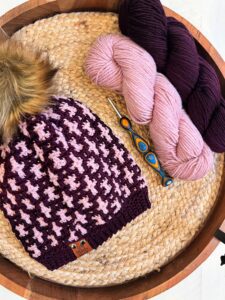 A mosaic crochet hat featuring a pink and maroon houndstooth pattern with a faux fur pom-pom is placed on a woven circular basket. Next to the hat are two skeins of yarn, one pink and one dark maroon, along with a crochet hook. -Marly Bird