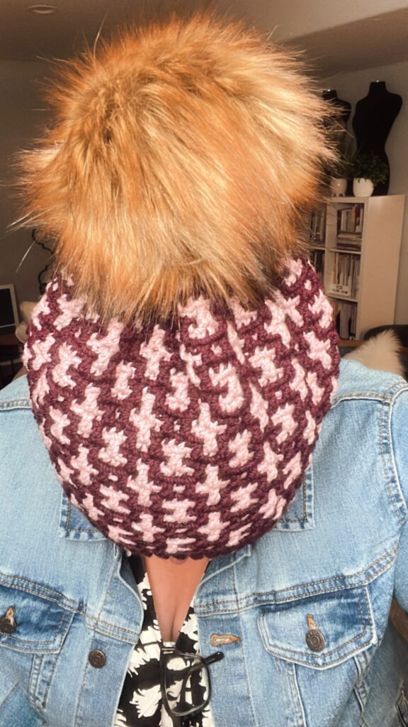 Person wearing a maroon and pink houndstooth patterned mosaic crochet hat with a large brown faux fur pom-pom, looking down. They have on a denim jacket and are indoors, with shelves and a couch visible in the background. -Marly Bird