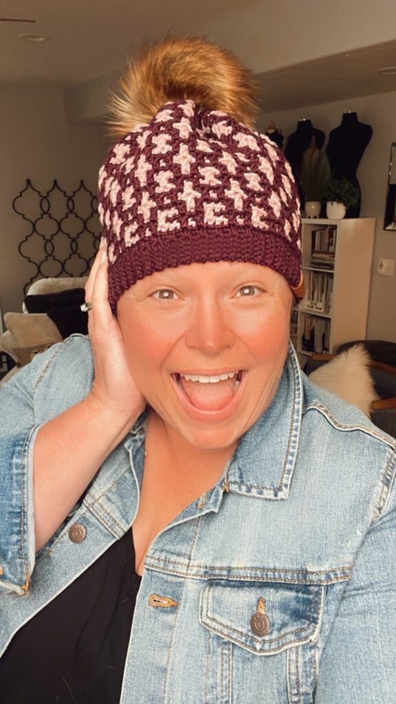 A person with short hair and a joyful expression poses indoors, wearing a maroon and white houndstooth knit beanie with a pom-pom on top. They are dressed in a denim jacket over a black shirt and have a hand resting on their cheek, resembling the cozy charm of a Mosaic Crochet Hat. -Marly Bird