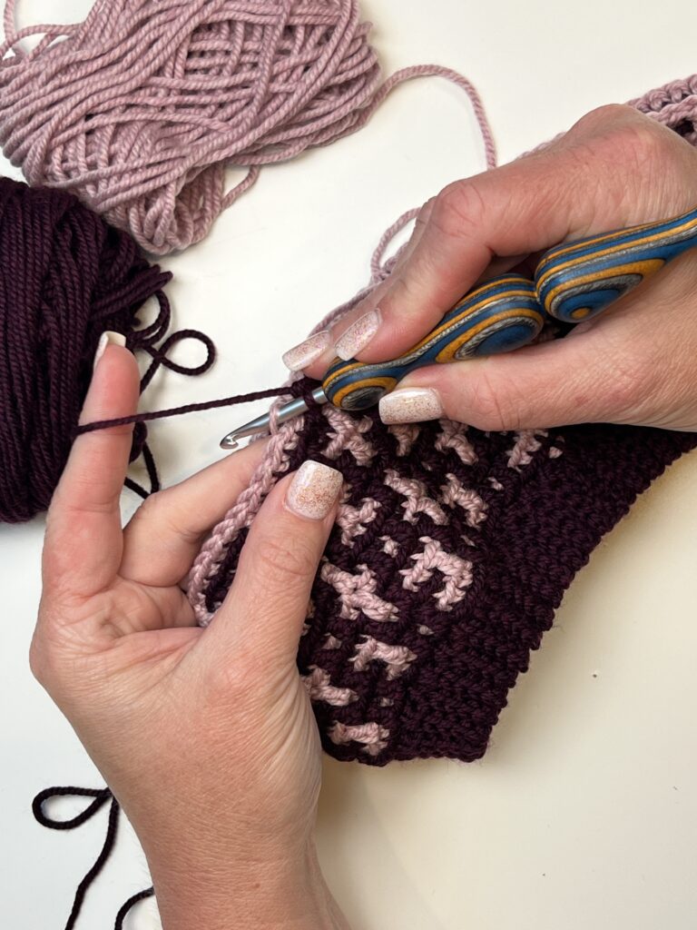 A person with manicured nails is crocheting a mosaic crochet hat with a colorful, ergonomic hook. They are working with dark purple and light pink yarns to create a textured pattern. Two balls of yarn are seen in the background on a white surface. -Marly Bird