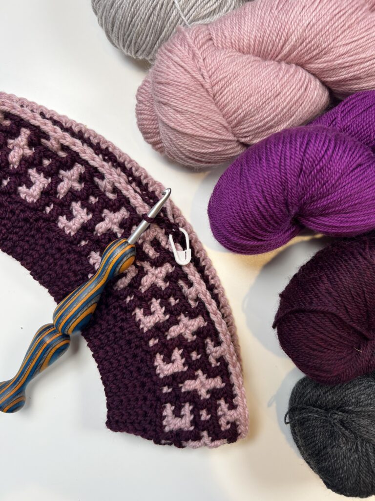 A crochet project in progress using pink and purple yarns is shown alongside skeins of gray, light pink, purple, and dark gray. The pattern includes a geometric design perfect for a mosaic crochet hat. The workpiece is paired with a wooden crochet hook with blue and brown stripes. -Marly Bird