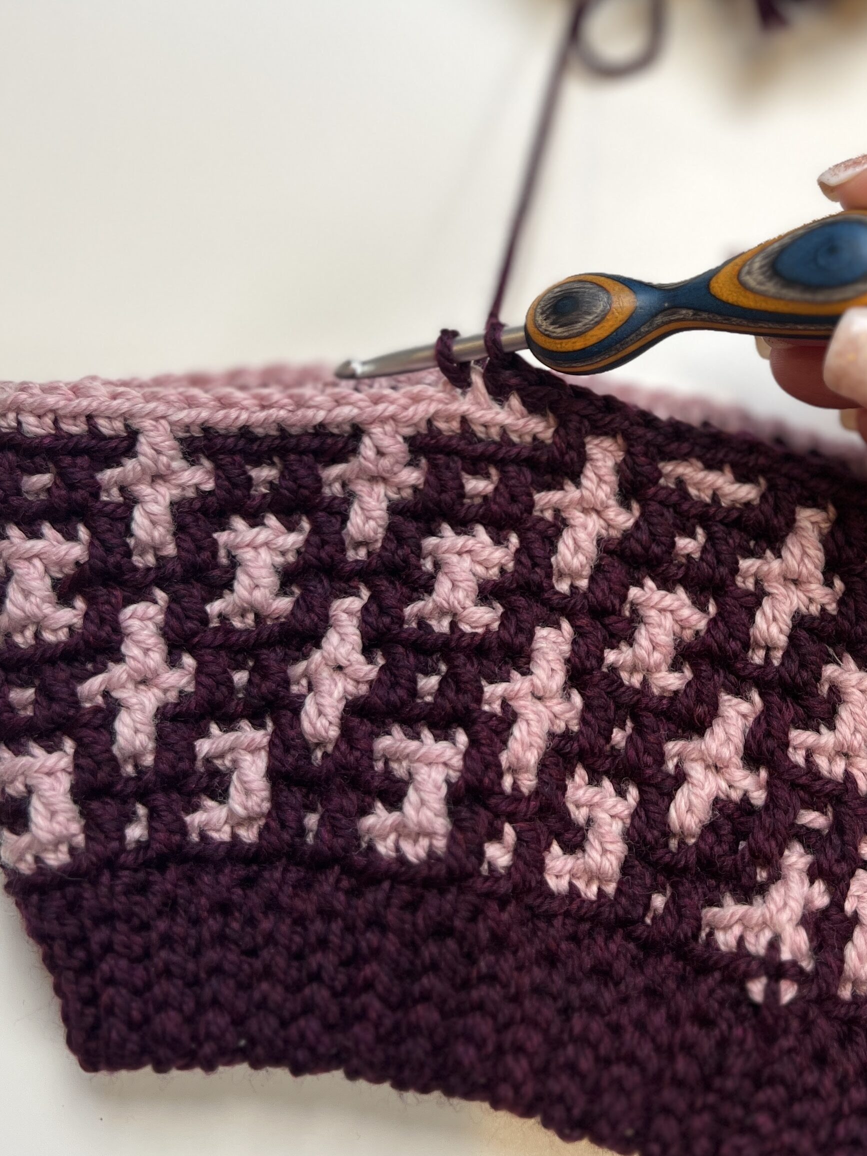 Close-up of a hand holding a crochet hook, working on a mosaic crochet hat with a purple and pink houndstooth pattern. A ball of purple yarn and a partial ball of pink yarn are in the background on a white surface. -Marly Bird