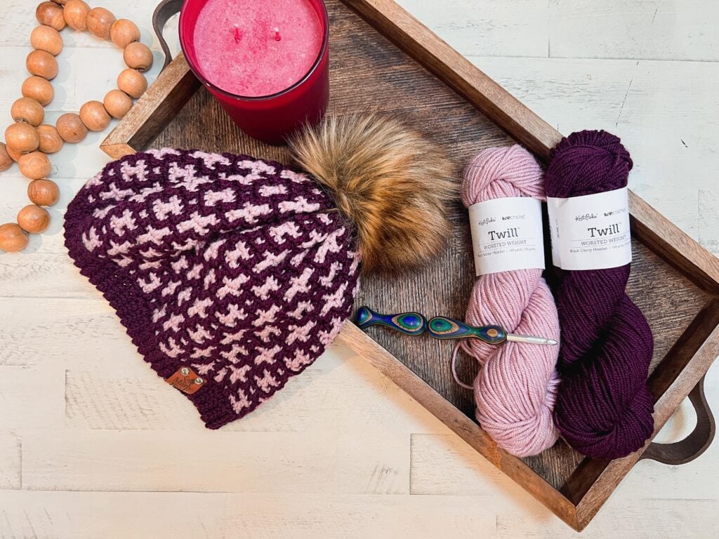A wooden tray holds a pink candle, wooden beads, two skeins of yarn, a crochet hook, and a knit hat with a faux fur pom-pom—perfect for those seeking inspiration or free knitting and crochet patterns in dark purple and light pink tones. -Marly Bird
