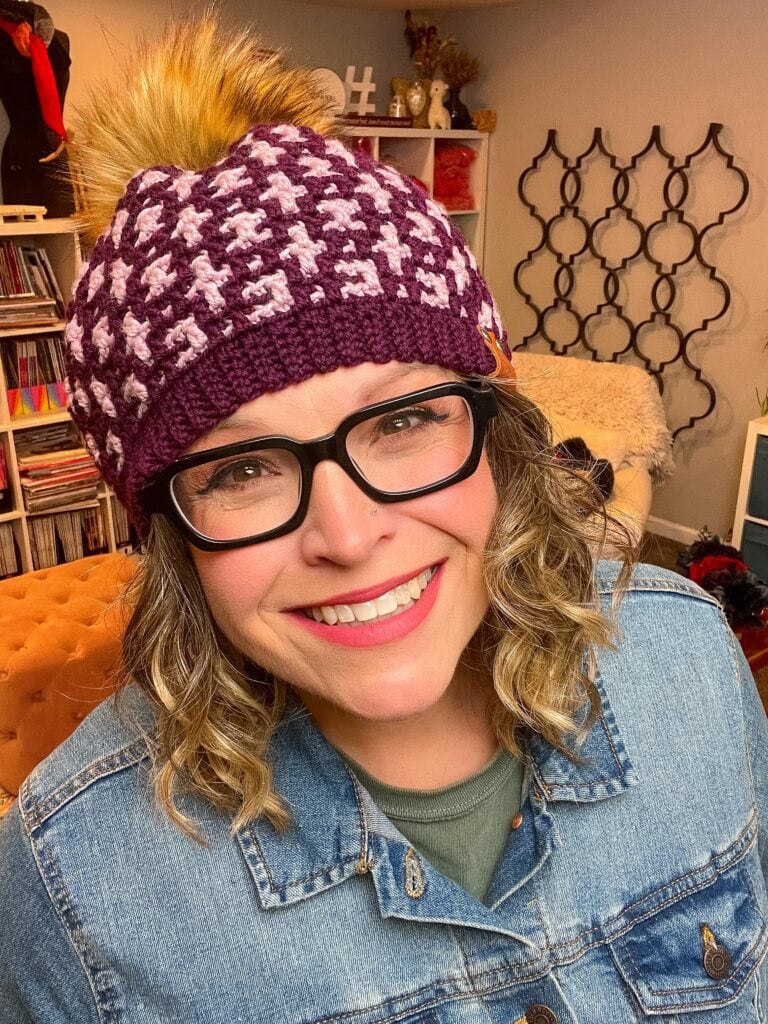 A smiling woman wearing glasses, a purple Mosaic Crochet Hat with a pom-pom, and a denim jacket stands indoors with shelves and patterned decor in the background. -Marly Bird