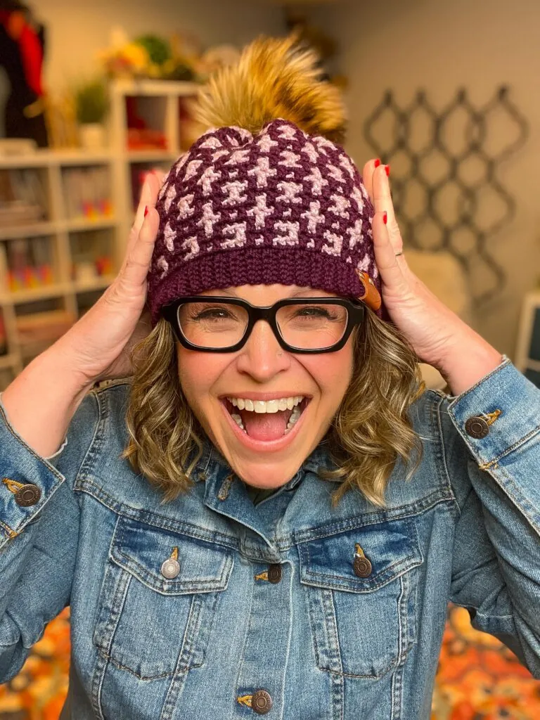 A smiling woman wearing glasses, a denim jacket, and a purple mosaic crochet hat with a pom-pom holds the sides of her hat, standing indoors with bookshelves and a patterned wall in the background. -Marly Bird
