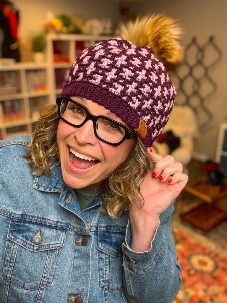 A smiling woman wearing glasses, a purple and white Mosaic Crochet Hat with a pom-pom, and a denim jacket poses indoors, holding the hat’s edge with one hand. Shelves and colorful decor are visible in the background. -Marly Bird