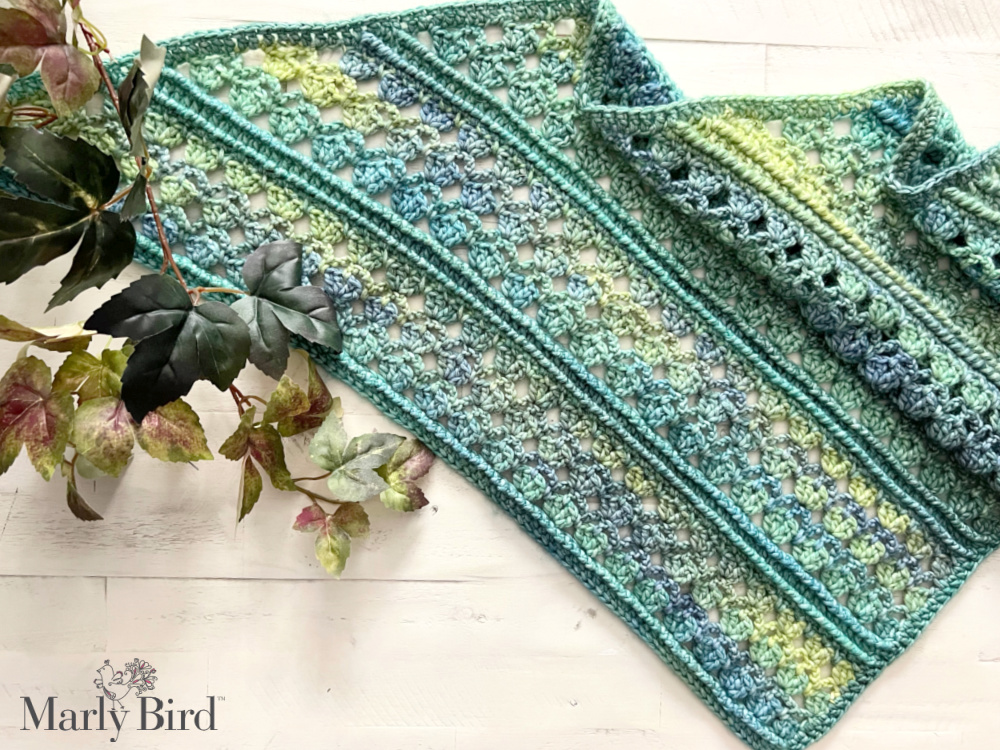 Mangrove Crochet Shawl Sample on white table. Shawl is in Tidepool colorway of Caron Blossom Cakes yarn - Marly Bird