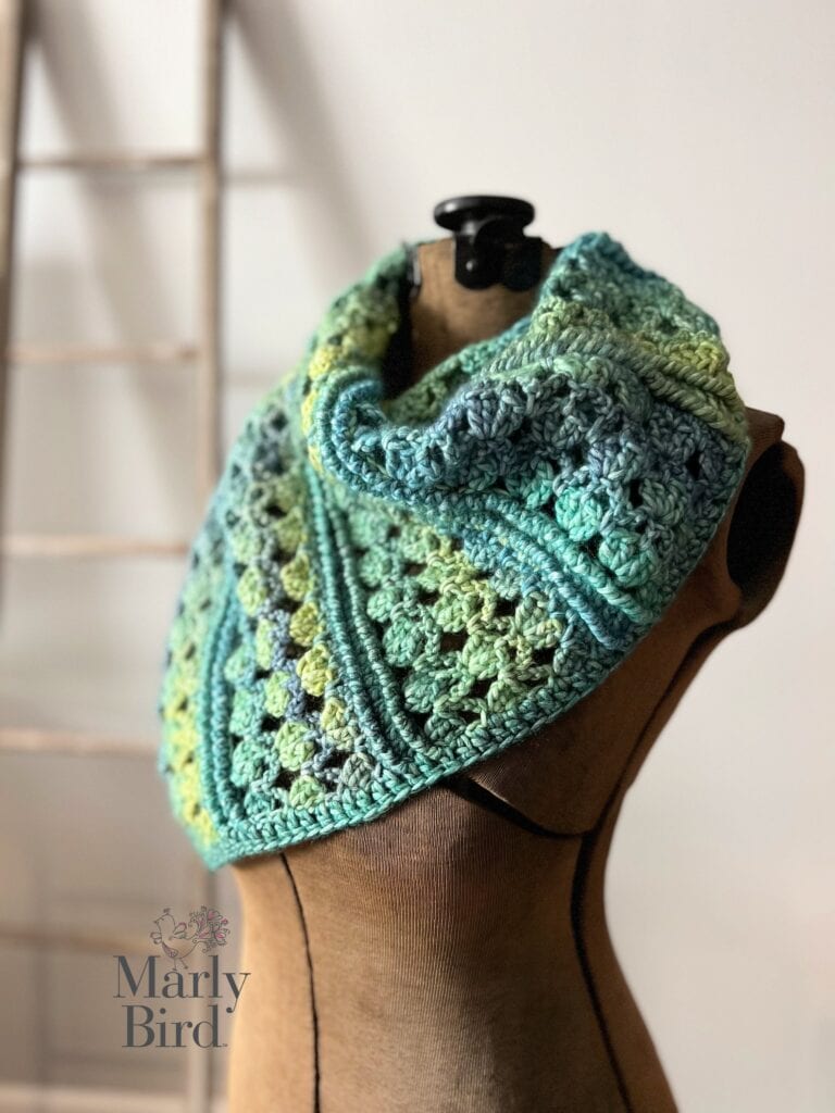 Crocheted cowl in blue and green tones displayed on dress form, showing textured stitches; blurred wood ladder background.