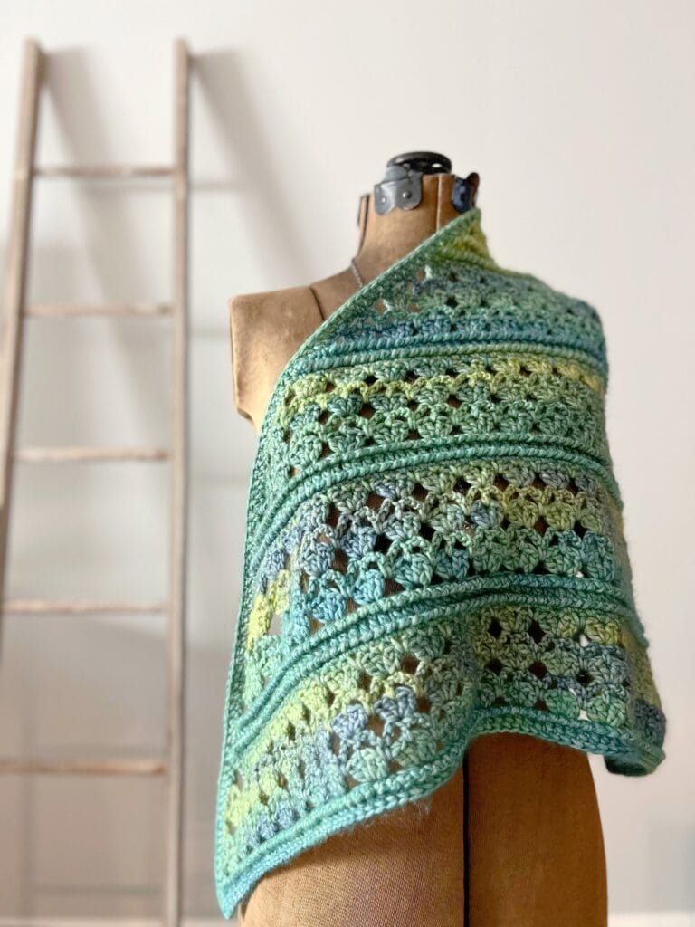 A dress form displays a blue and green crocheted shawl with an intricate pattern, perfect for those seeking free knitting and crochet patterns. In the blurred background, a wooden ladder leans against a plain white wall. -Marly Bird
