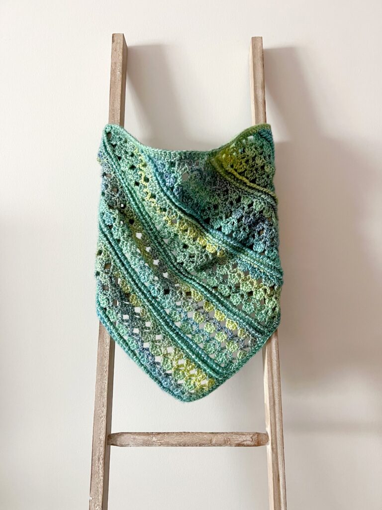 A crocheted shawl in green, yellow, and blue tones hangs on a wooden ladder against a plain white wall. The shawl features textured and openwork patterns, perfect for those looking for inspiration or free knitting and crochet patterns. -Marly Bird