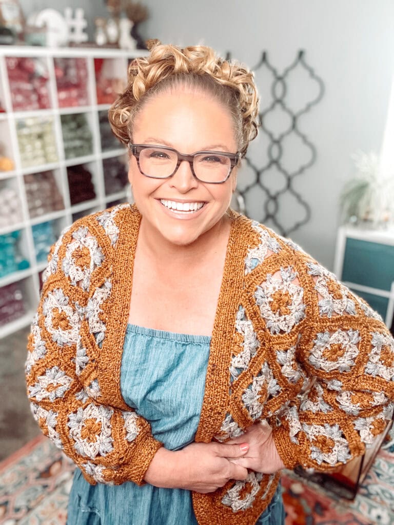 A smiling woman wearing glasses, a crocheted granny square cardigan, and a blue dress stands indoors with shelves of yarn in the background. The scene is bright and cheerful. -Marly Bird
