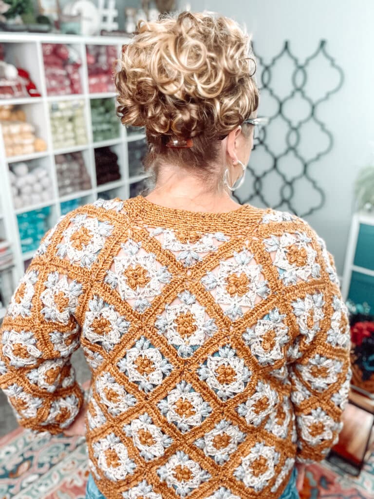 A woman with curly hair in a bun, wearing glasses and a crocheted sweater with a yellow and white granny square pattern, stands facing a yarn storage shelf filled with colorful yarn. -Marly Bird