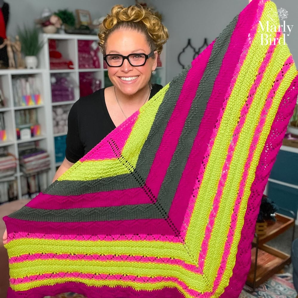 A woman displays a knit shawl in pink, yellow, and gray stripes; textured worsted yarn. Yarn shelves appear in the background.